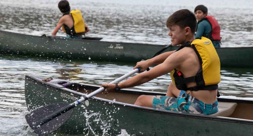 in the foreground, a boy paddles a canoe while two other boys paddle a canoe in the background